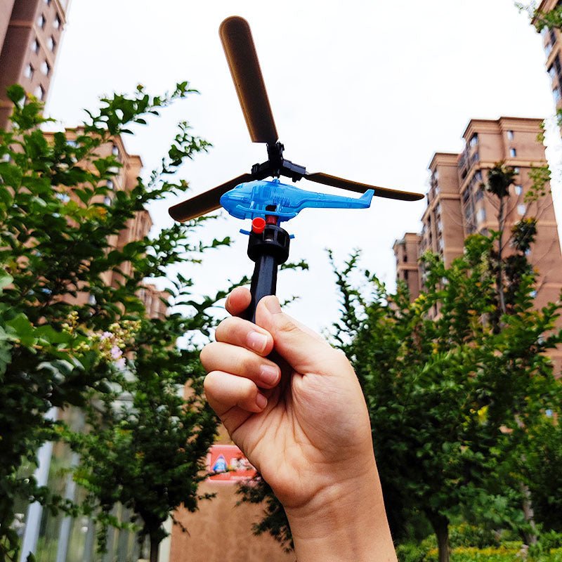 Hand holding blue pull-string helicopter toy launcher outdoors against trees and buildings